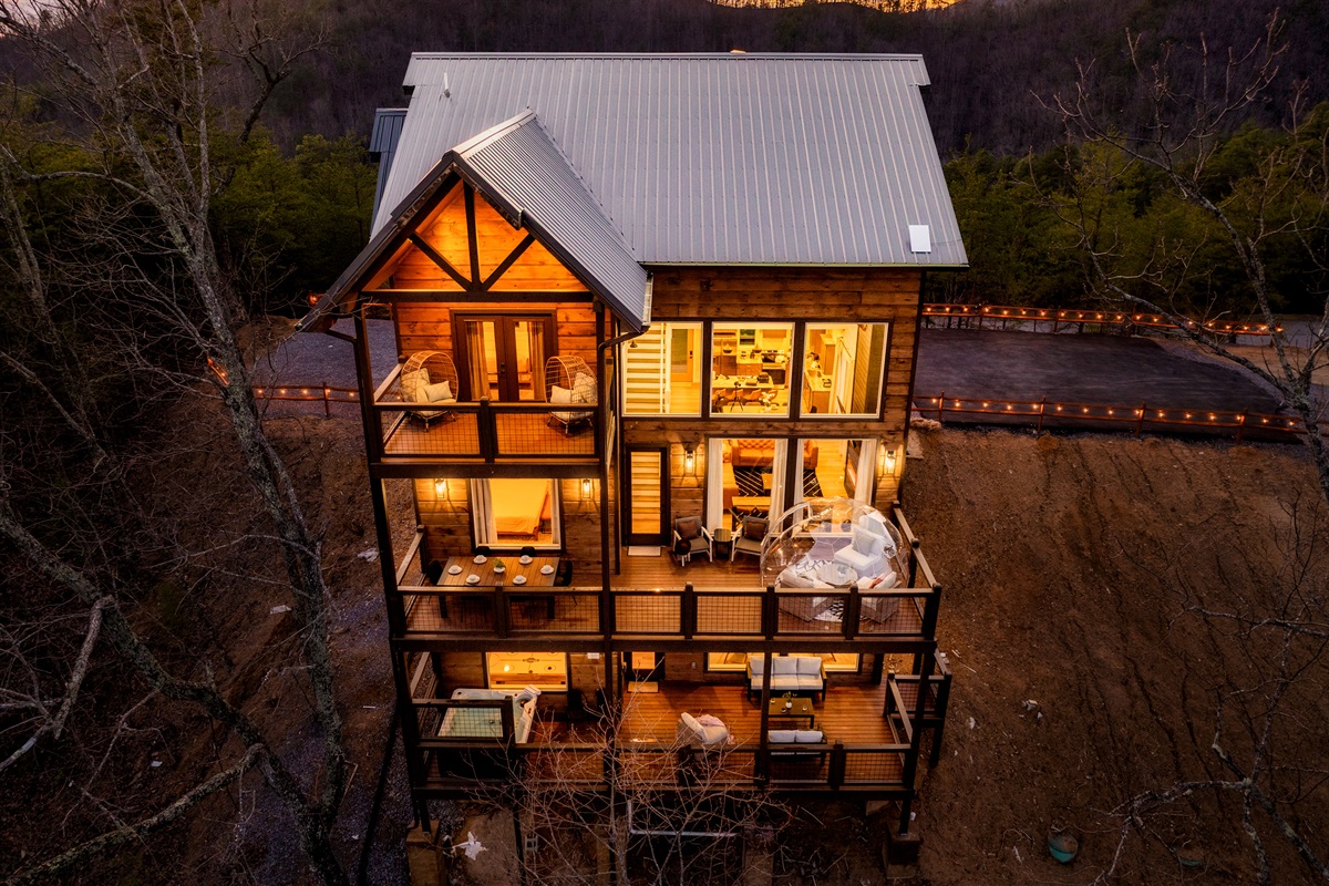 Aerial view of a modern wooden cabin with multiple levels, showcasing large windows, balconies with seating, and warm lighting against a backdrop of trees and a hillside.