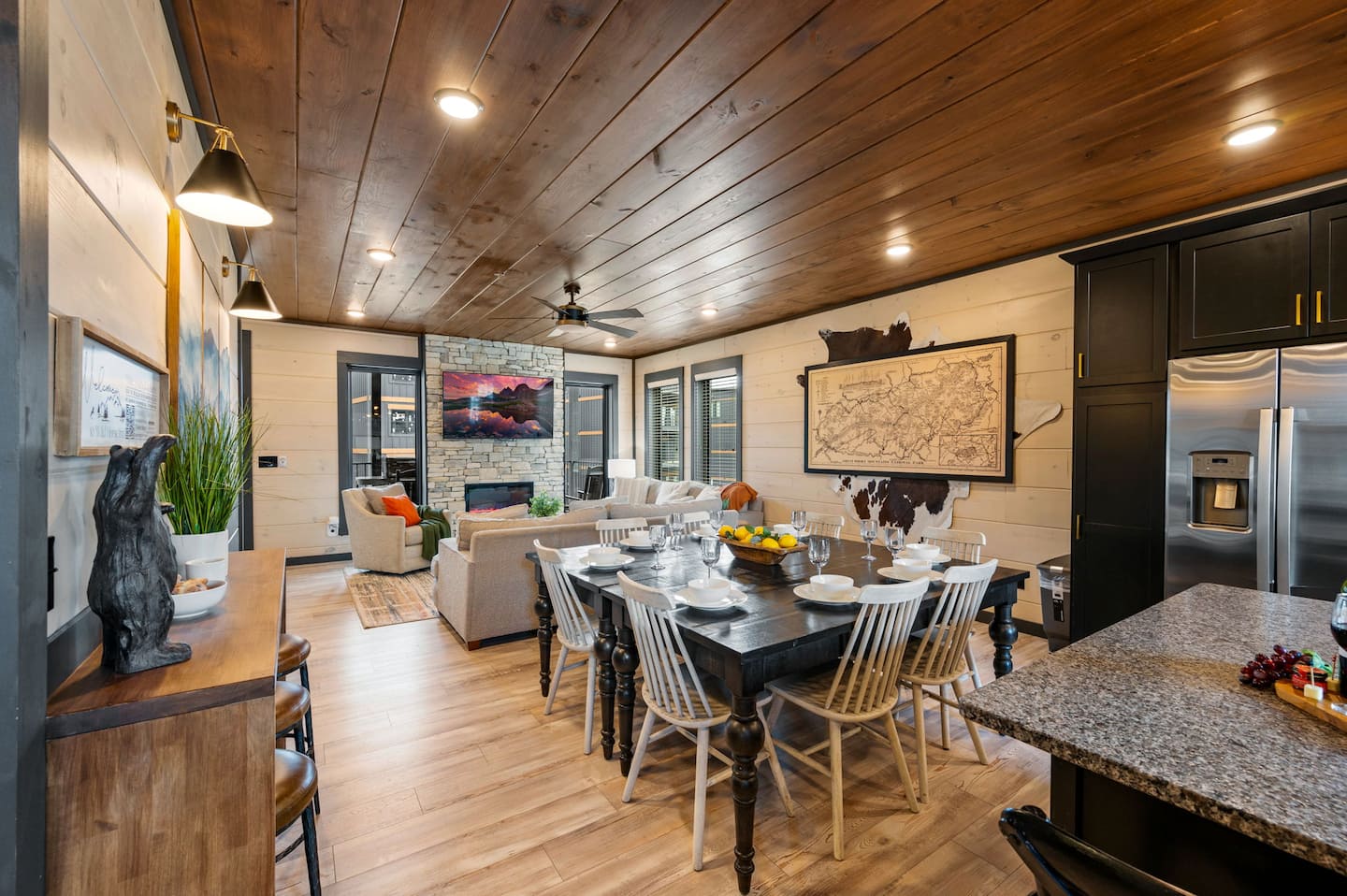 An inviting dining area with a large table set for meals, surrounded by wooden chairs, and a cozy living space featuring a stone fireplace, artwork on the walls, and natural light filtering through windows.