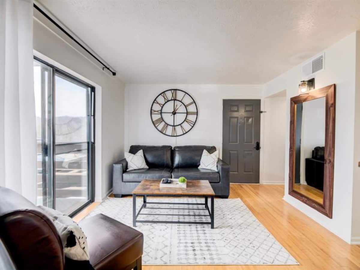 A cozy living room featuring a dark sofa with decorative pillows, a wooden coffee table, and a large wall clock. Natural light fills the space through a nearby window, and there is a mirror and a door visible in the background.