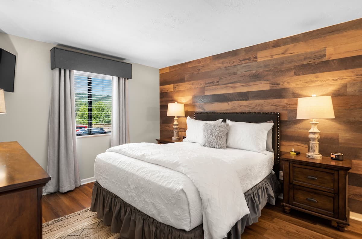 Cozy bedroom with a queen-sized bed dressed in white linens, wooden accent wall, lamp on nightstand, and window with light curtains.