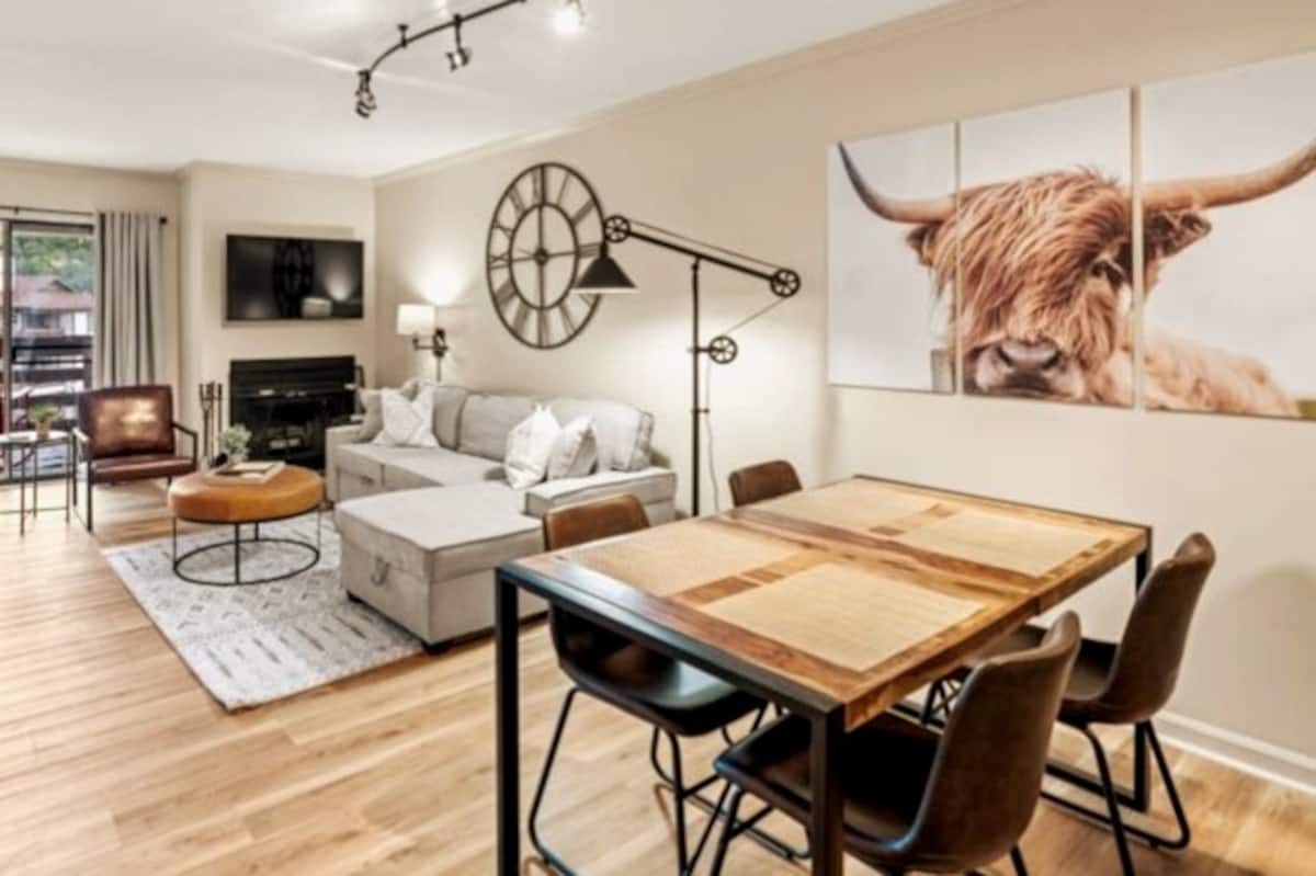 A cozy living area featuring a gray sectional sofa, a decorative wooden coffee table, and a dining table with black chairs. A large wall clock and a triptych artwork of a cow adorn the walls, while a sleek floor lamp provides warm lighting.